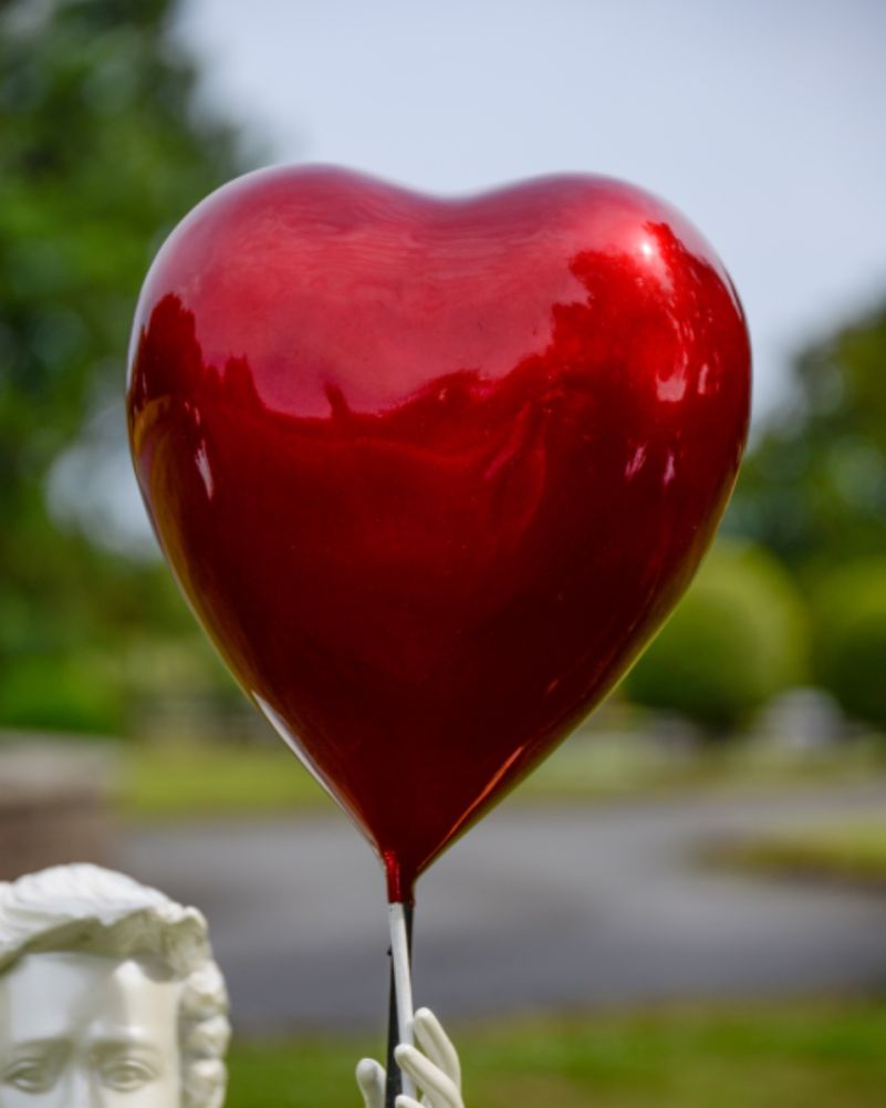 Close-up of the glossy, reflective metallic red heart-shaped balloon that the white statue of a girl is reaching to hold